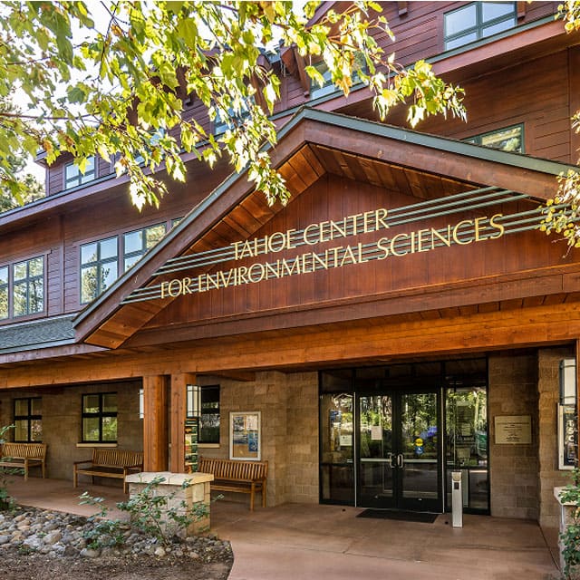 Entrance of the Tahoe Center for Environmental Sciences at the University of Nevada, Reno Lake Tahoe campus. The building features a rustic design with wood and stone elements, a peaked roof, large windows, and benches along the walkway shaded by leafy trees.