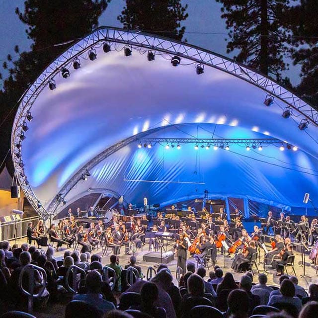 Evening outdoor concert at the Lake Tahoe campus featuring a full orchestra performing under a large white canopy with blue stage lighting, surrounded by pine trees and an audience seated in front.