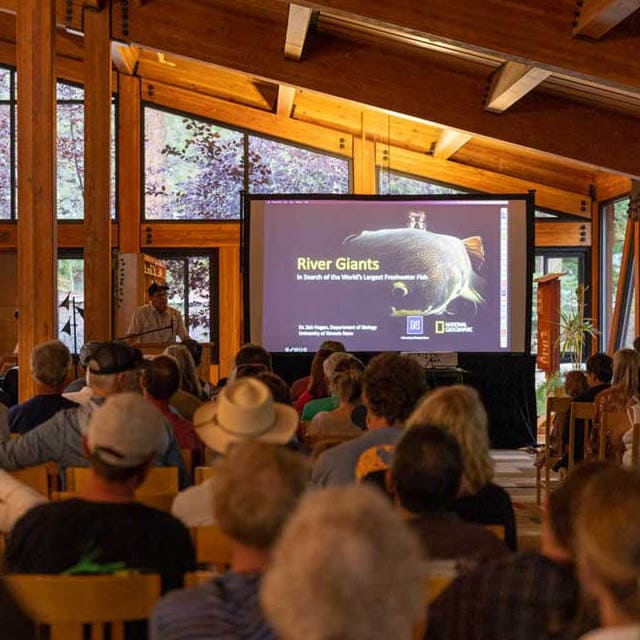 Indoor lecture at the Lake Tahoe campus with attendees seated in wooden chairs, listening to a speaker presenting a slide titled ‘River Giants’ on a large screen inside a wood-paneled room with tall windows.