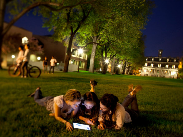 Students laying on the quad at night looking at an electronic device