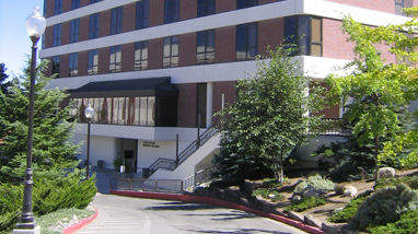 Exterior of the Ansari Business Building, a multi-story brick building with lined concrete white strips, and large green trees in front of the building.