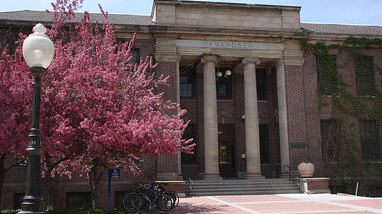 An exterior of Frandsen Building, a classic brick building with white pillars, large walking paths and purple trees in front of the building.