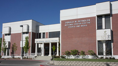 Exterior of the National Judicial building, an older brick building with white lined concrete roofing, bushes in front and the building's name on the side of the entrance.