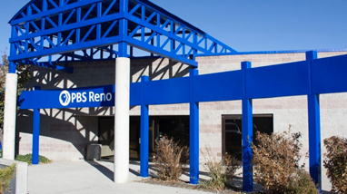 Exterior of the PBS Reno building, a concrete building with blue steel beams surrounding the building and the words "PBS Reno" and logo on the beam in front of the door.