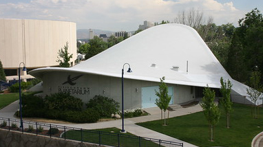 An exterior of the Fleischmann Planetarium and Science Center, a building with a parabolic white roof, walkways and trees surrounding the building.