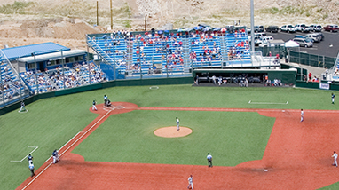 Spectators sit in blue stands at the William Peccole Park, a baseball field with green infield and dirt base paths, on the campus of the University of Nevada, Reno.