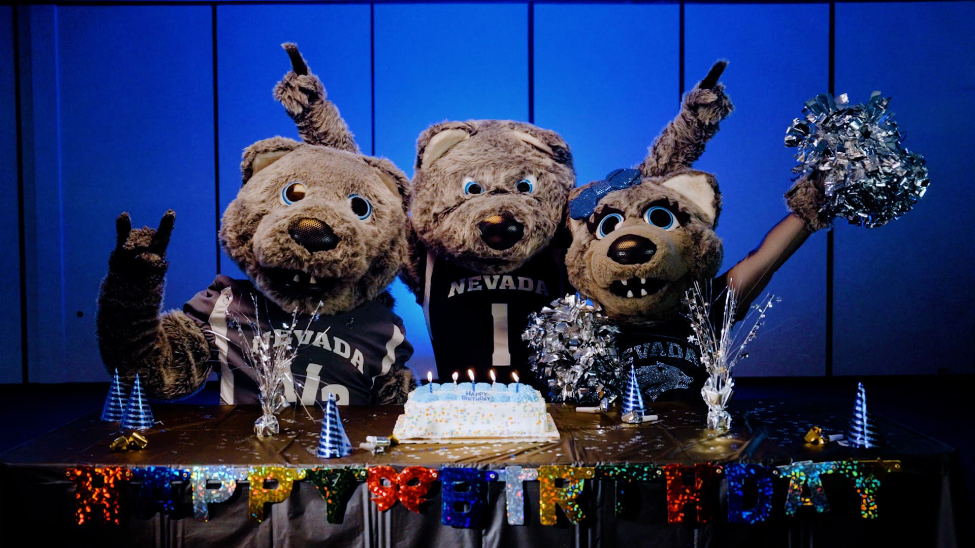 University mascots, Alphie, Luna, and Wolfie Jr., wear Nevada jerseys and celebrate around a birthday cake.
